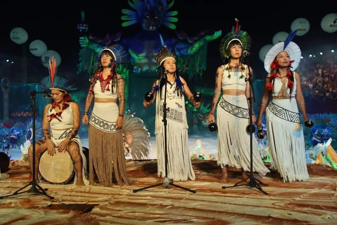 As Karuana se apresentando ao vivo no palco, cinco integrantes com adornos indígenas tradicionais